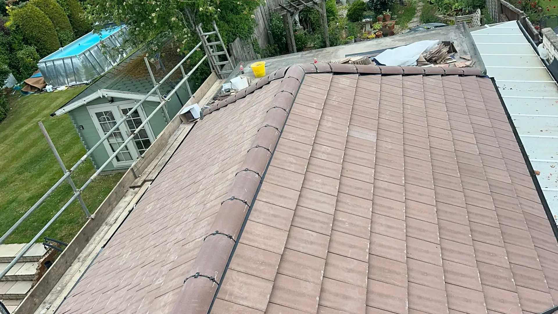 Newly re-roofed hip roof in brown plain tiles with a new ridge, scaffolding and a garden below